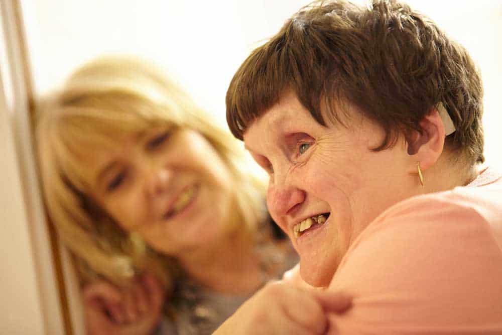 A woman in a pink t shirt looks to the right. In the background, a woman is smiling at her