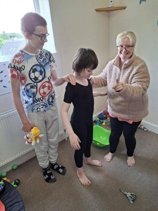 Two boys and a woman are in a room. The boys are looking at the floor, their mum is pointing to the camera. One boy is wearing a black leotard.