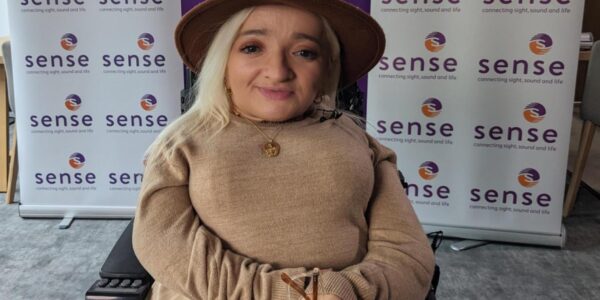 A stylishly dressed woman wearing a hat sits in her wheelchair in front of banners with the Sense logos printed in a grid formation.