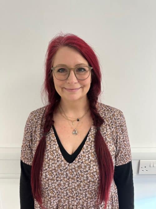 A woman with long dark pink hair tied in two plaits smile to the camera.