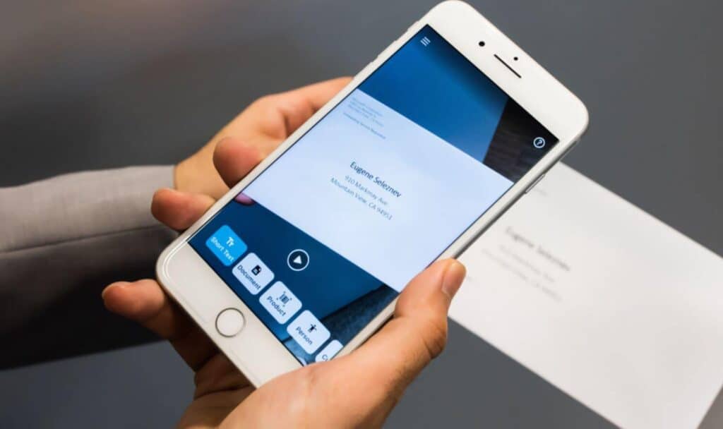 A close-up of a hand holding a smartphone, which is open to the camera in the Seeing AI app, pointing at an address on an envelope.