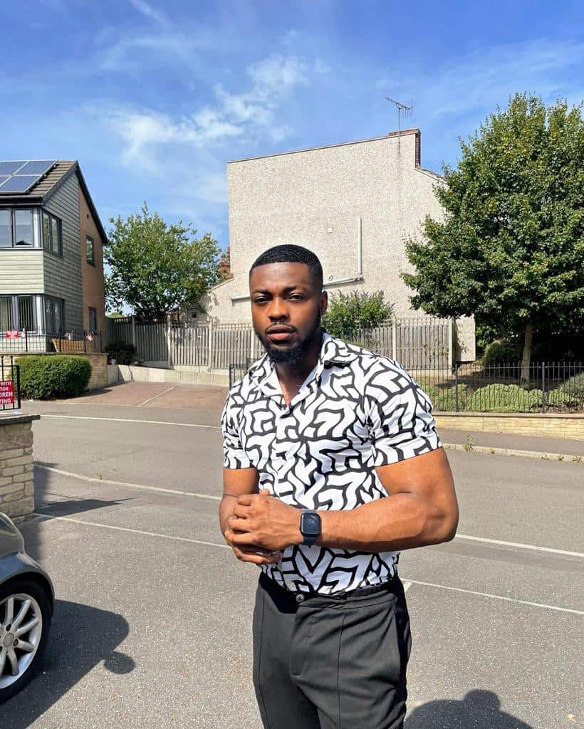 A young man in a short sleeved shirt stands on a sunny roadside.