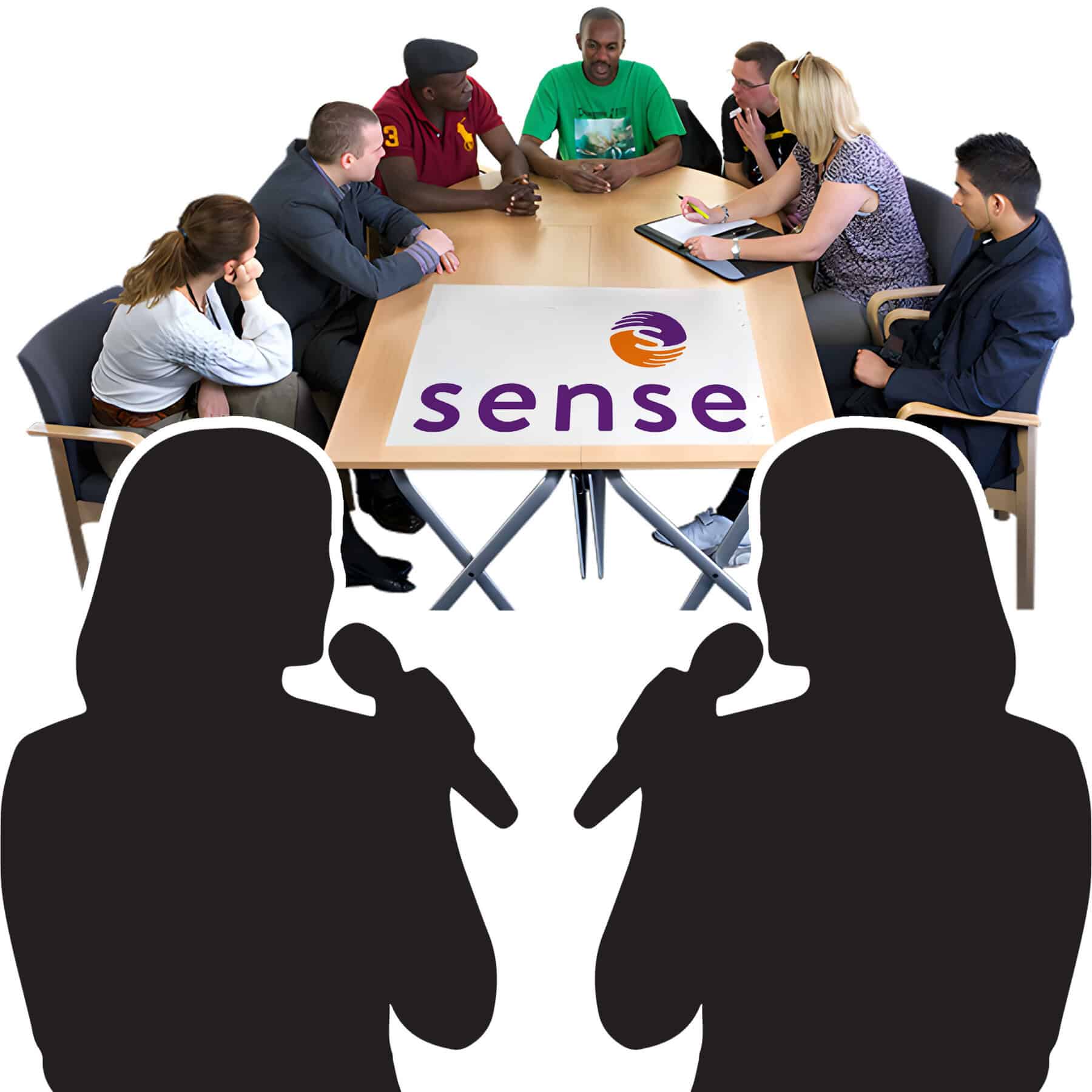 A group of people discuss things sitting around a table. 2 people stand in front of them talking into microphones