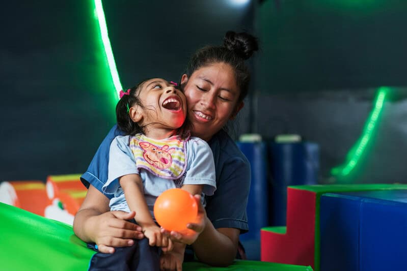 A woman stands behind a smiling girl holds an orange ball out in front of her.