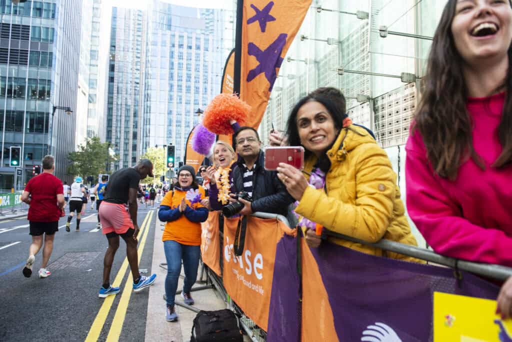 A cheering woman leans on a fence post watching marathon runners pass through the streets of London.