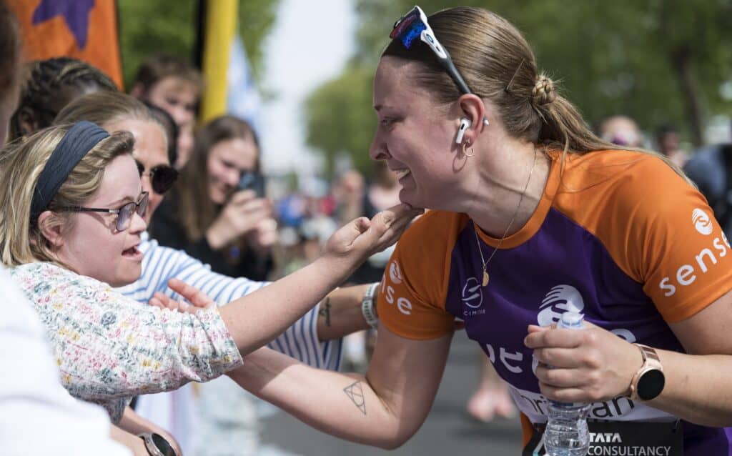 A sense marathon runner and her sister share an emotional moment on the side of the track. The sister is lifting up the chin of the runner as they talk.