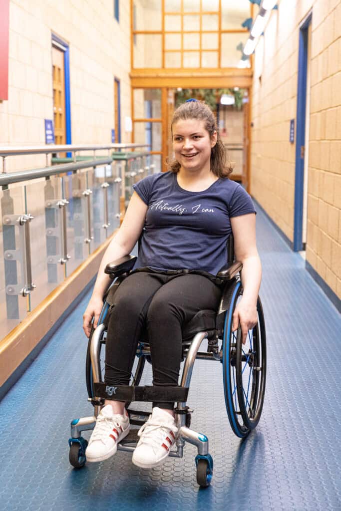 A young white woman in a wheelchair in the corridor of her Sense College.