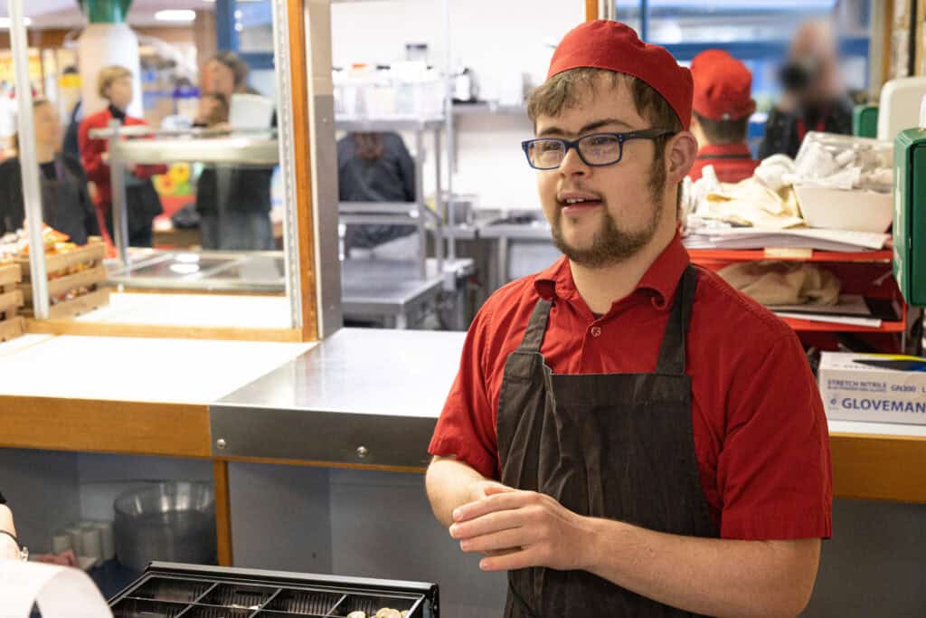 A man in a hat and apron looks to someone off camera that he's serving in a café