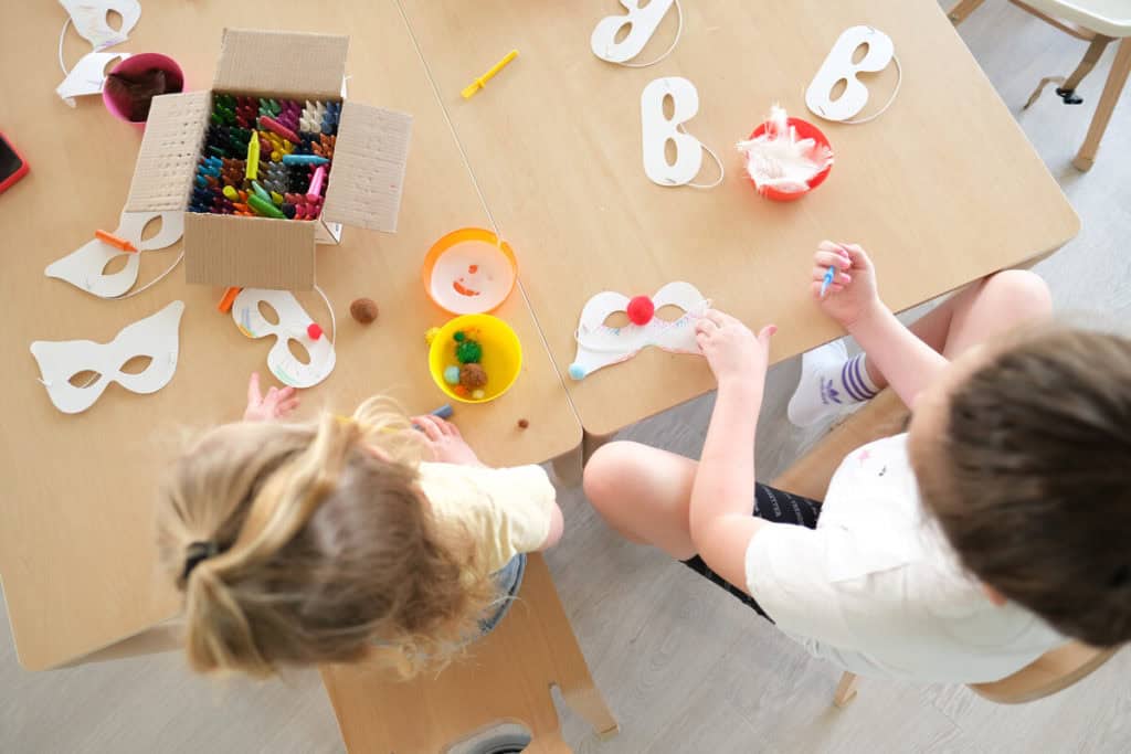 Two children photographed from above, sitting at a table filled with masks and arts and crafts supplies