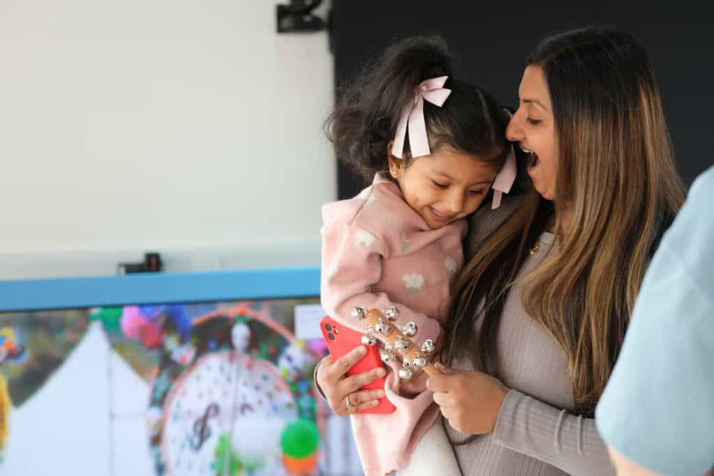 A little girl with ribbons in her hair laughs while being held by an adult who is holding a wooden musical instrument covered in bells.