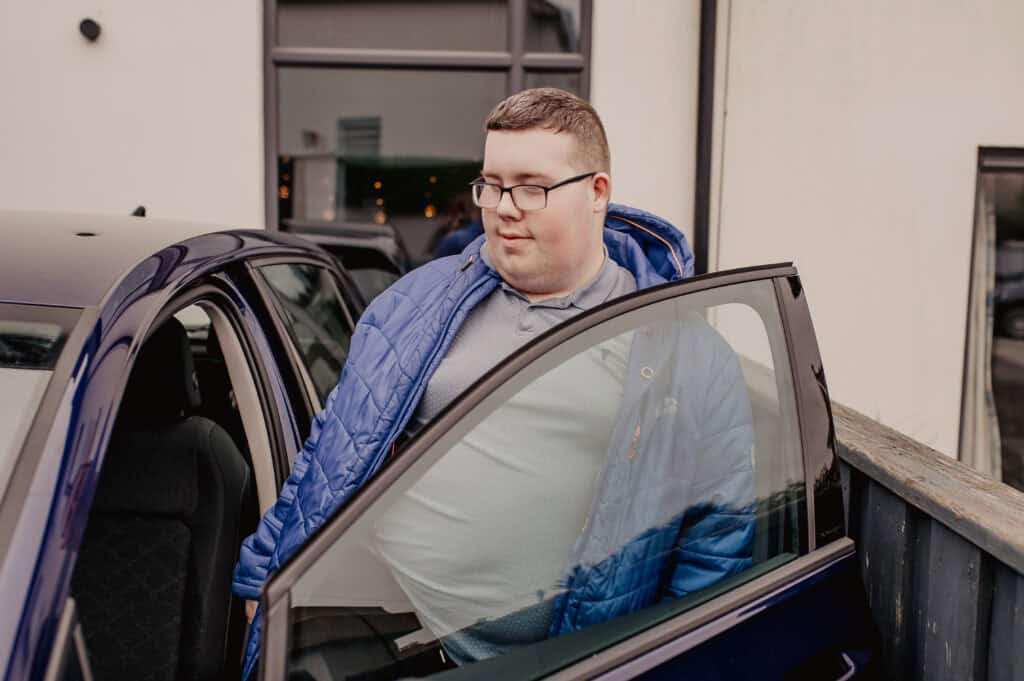 A young man wearing glasses gets into a blue car.