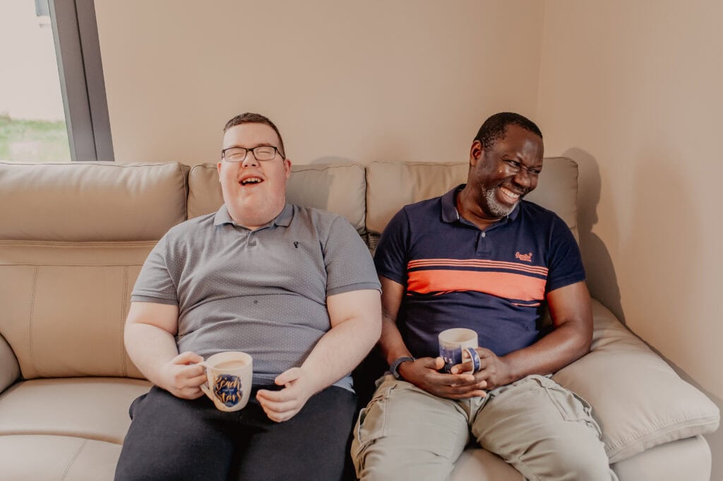 Jamie sharing a laugh with a support worker as they both drink a cup of tea on the sofa.