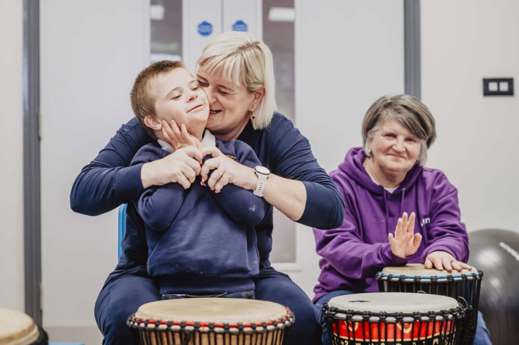 A mother has her arms wrapped around her son, he is smiling into her cheek. In front of them there are bongo drums.