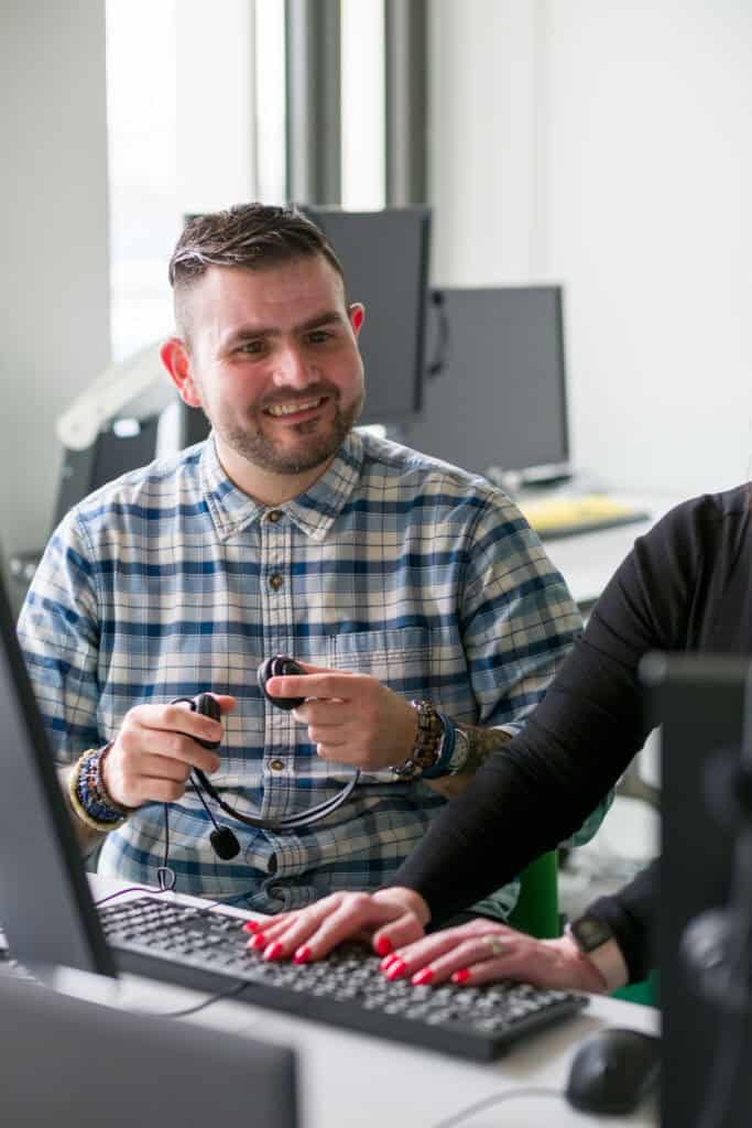 Chris, a smiling man wearing a checked shirt, holding a headset in front of a computer.