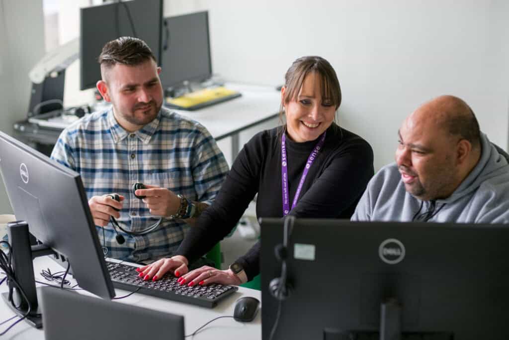 Two men sit either side of a woman at a computer in an office, all laughing and chatting together.