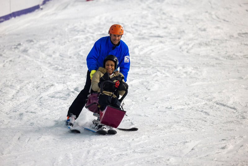 A woman on a sit ski is guided down a snowy slope by her instructor, who's skiing close behind her.
