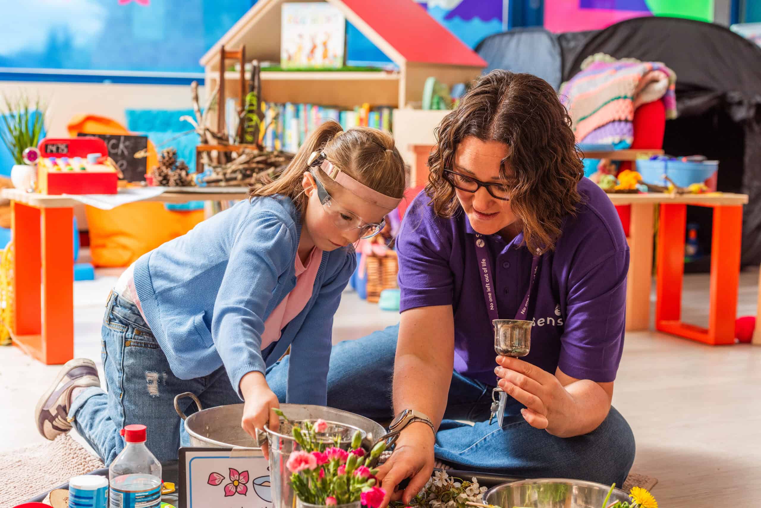 A young girl in a blue cardigan is leaning over to grab a bucket of flowers, a lady with dark hair is sat next to her