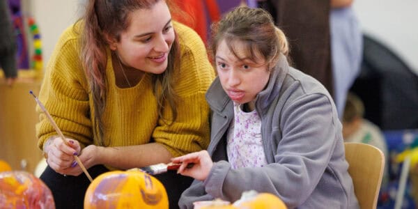 A girl is supported by a smiling support worker during a pumpkin-painting activity.