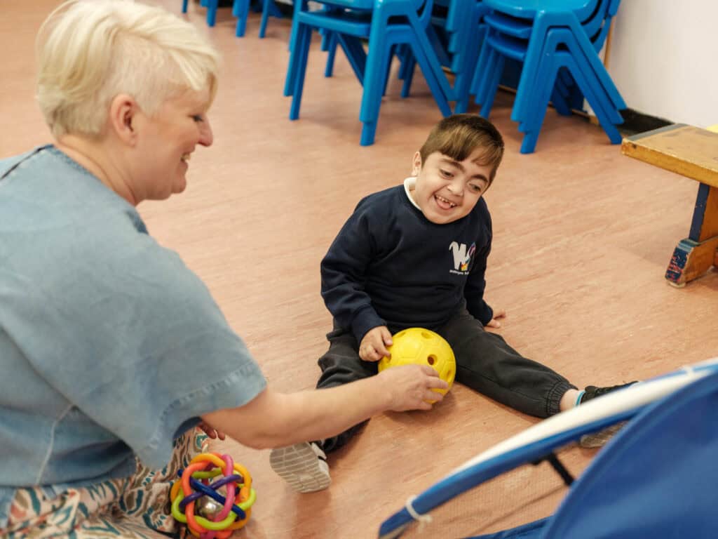 Amin laughs while playing with a yellow ball with his support worker, an adult woman with short blonde hair.