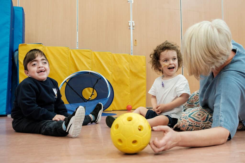 Two small children, Amin and his friend, laughing as they play sensory football with an adult support worker.