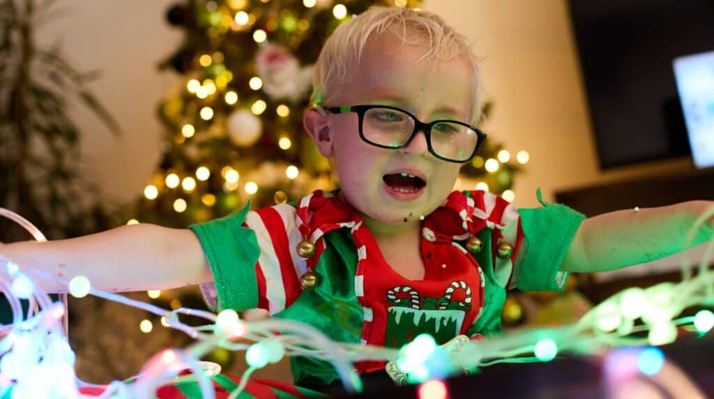 Harvey is sat in his elf suit, with his glasses on. He is playing with a string of green fairy lights in front of him.