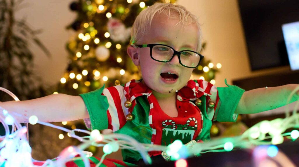 Harvey is wearing glasses and sat in his elf suit, inside a messy play area. He is smiling widely back at the Sense employee.
