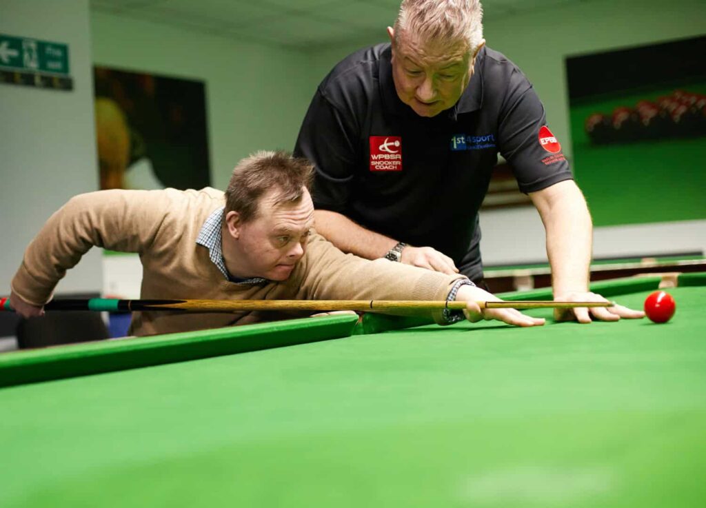 A man leans over a snooker table aiming his cue at a ball, an instructor is over his shoulder helping to guide the shot