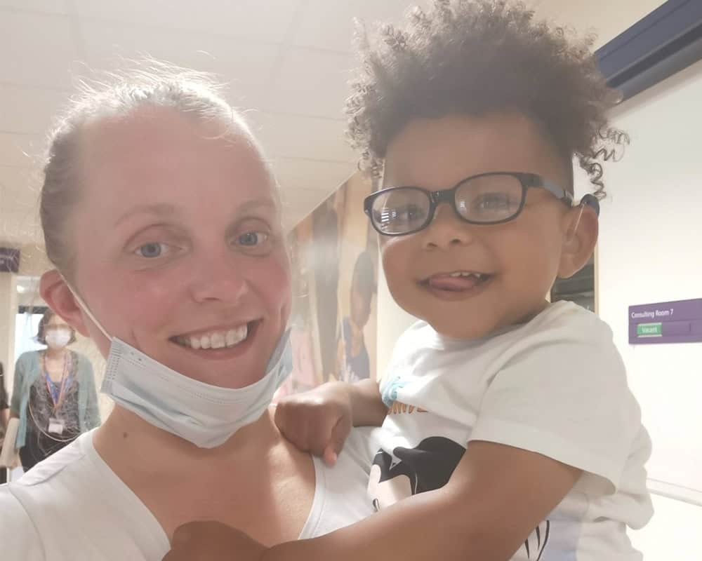 Simona holds Leo - she has blond hair and is wearing a surgical mask under her chin and Leo has curly hair and is wearing glasses. They are both smiling at the camera.