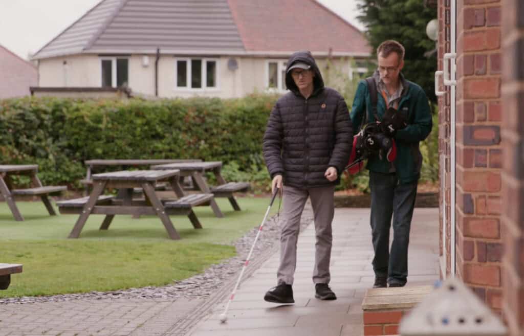Simon walks towards the camera using a white cane to identify the path. Behind him, another man carries Simon's golf clubs.