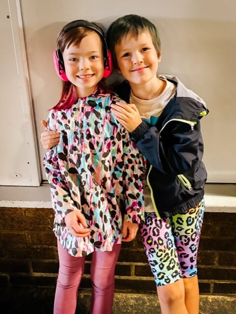 Two children, a boy and a girl. The boy has his arm around the girl, who is wearing ear defenders.