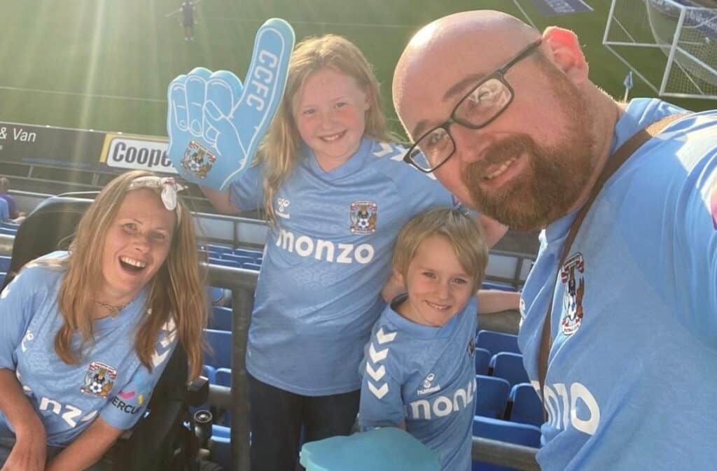Steph and her family pose for a picture inside Coventry Cities football stadium. They are all smiling widely, wearing match shirts with the daughter waving a light blue foam hand.