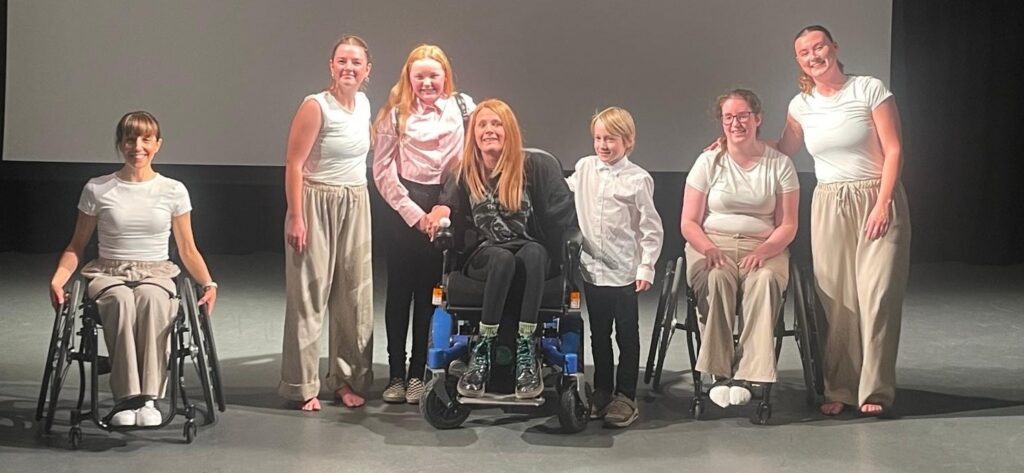 Steph is sat in her wheelchair, on a stage, surrounded by fellow dancers and her children.