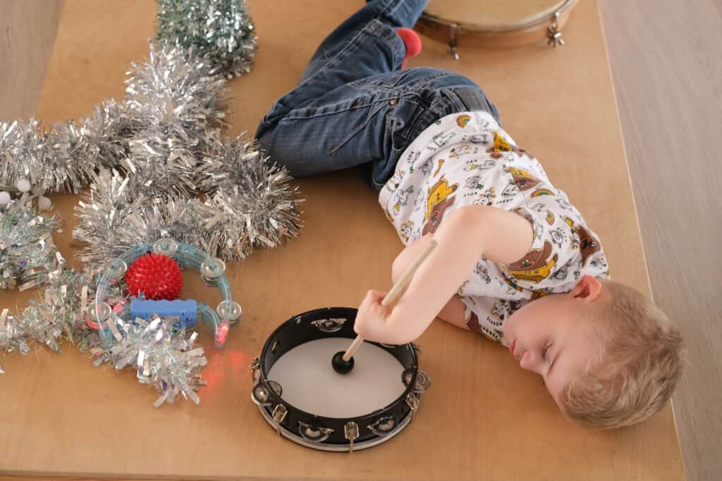 Thomas, a young boy, lying on his side on a resonance board with a tambourine, drumstick and tinsel.