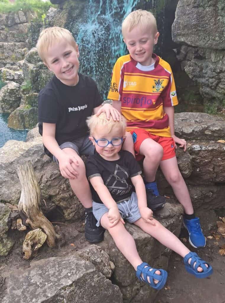 Three boys by a waterfall. One boy has his hand on top of the youngest one’s head.