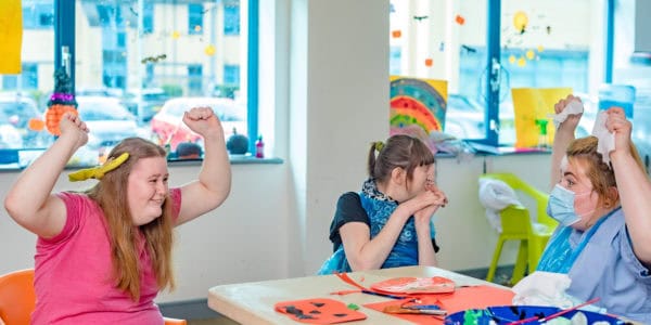 A young woman and support worker wearing a mask both raising their arms and smiling at an arts and crafts table.