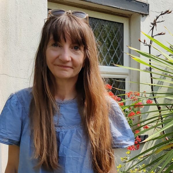 A white woman with long brown hair stands in front of a white building. She's wearing a blue blouse and has sunglasses on her head. Behind her, there's a white building that looks like a cottage and there's a ledge with some orange flowers on. Just behind her, there are big pointy green leaves coming from a bush that we can't see.