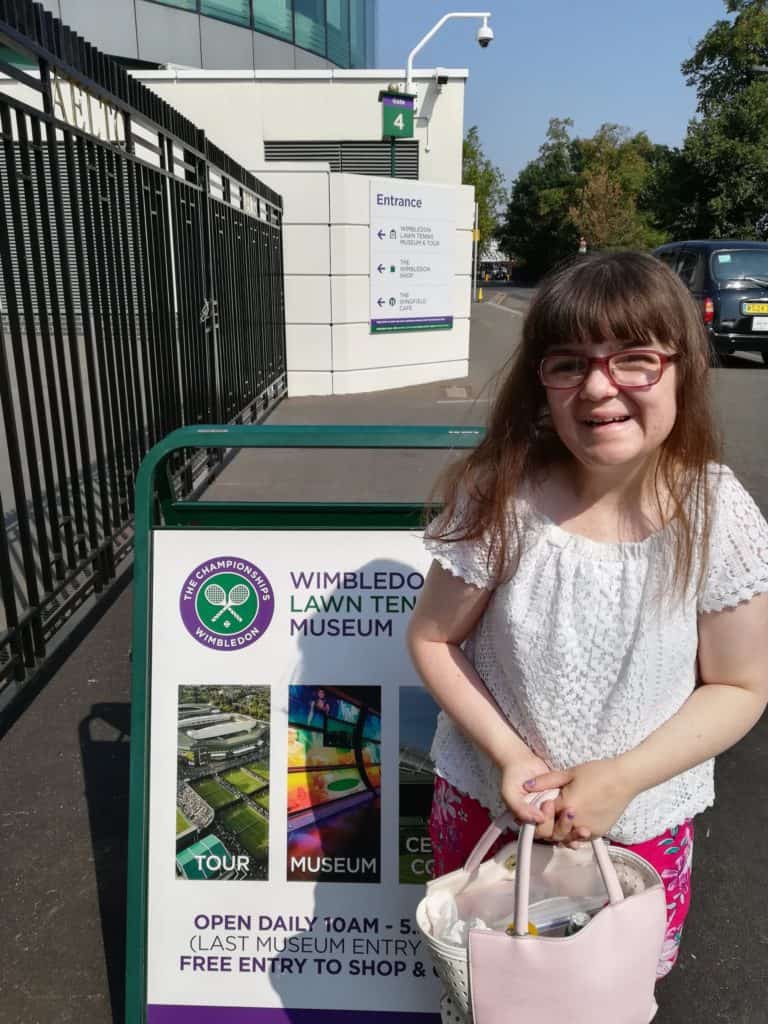 Sian, a young woman with long brown hair, stands smiling in front of a Wimbledon Lawn Tennis Museum sign.