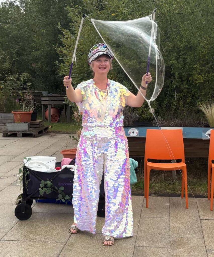A volunteer dressed in a sparkly sequin suit and tiara, is using a handheld bubble making device to make a huge bubble.