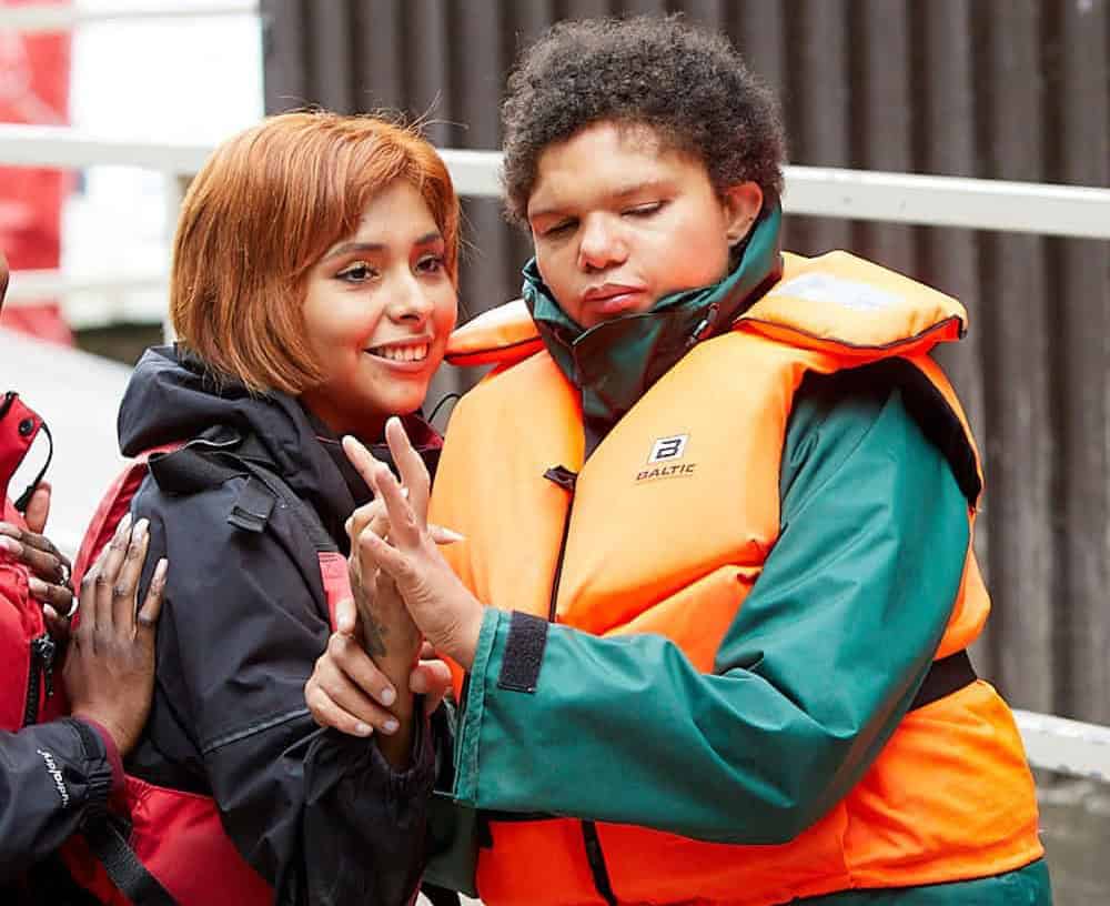 Two young women are stood side by side, touching hands and smiling. They are both wearing life jackets and rain coats.