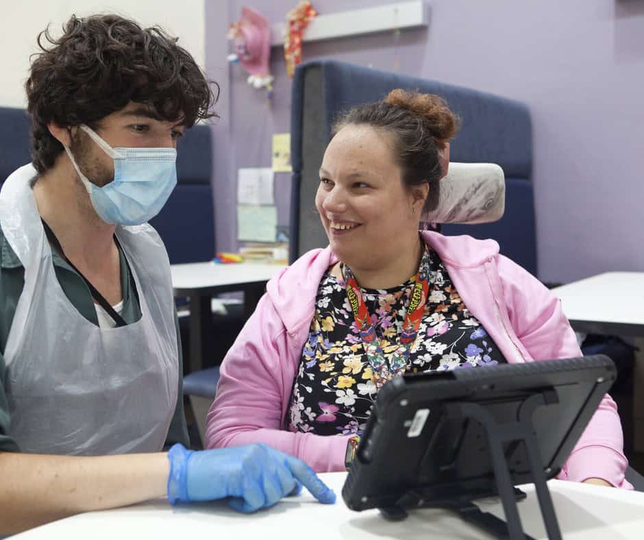 Maria, a white woman with brown hair in a bun is smiling at a man in PPE. She is wearing a pink hoodie and has a lanyard. They are looking at an iPad