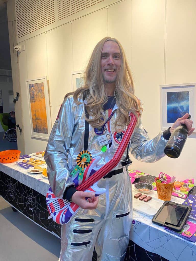 A man in a fancy dress costume wearing a long blonde wig and a silver space suit, holding an inflatable guitar.