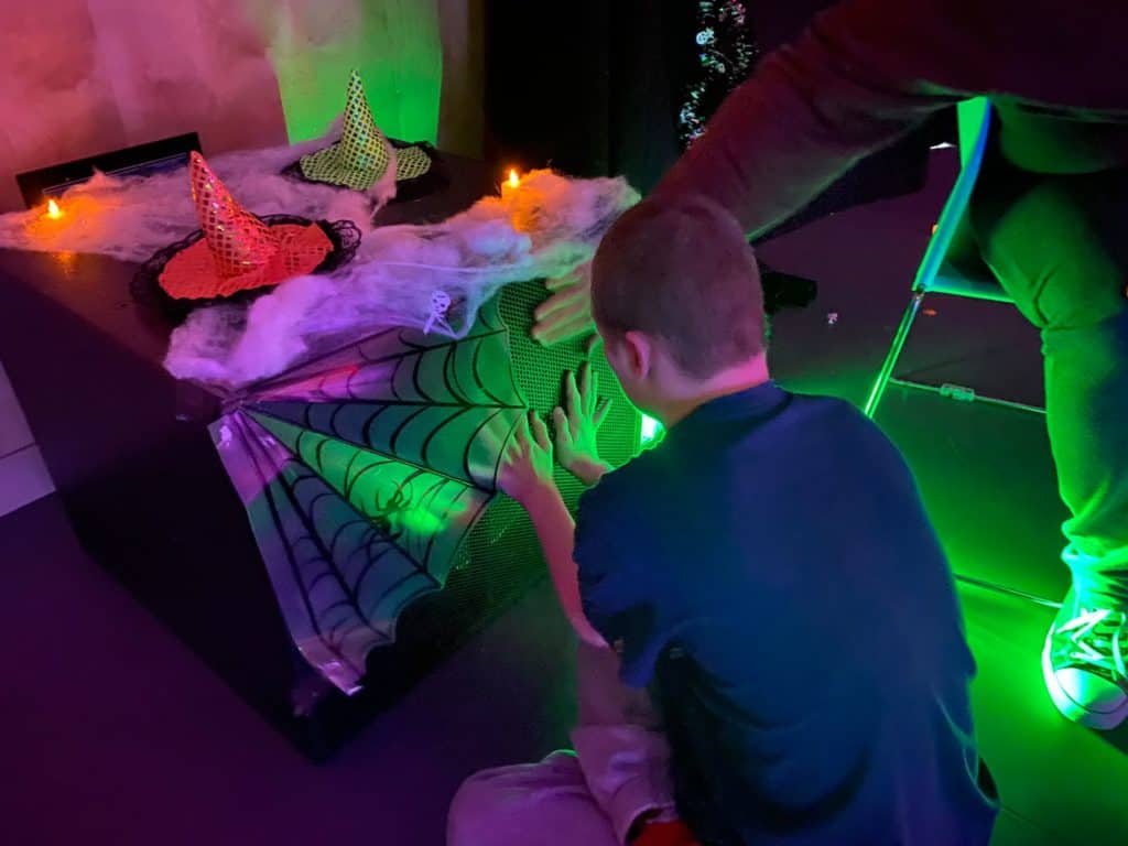 A young man places his hands on a speaker to feel the vibrations of the music being played. The speaker is decorated for Halloween with cobwebs.