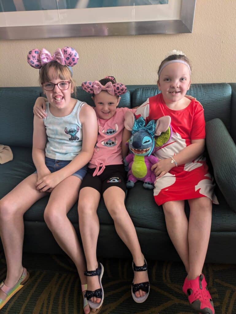 Three girls sitting on a sofa. Two are wearing Mickey Mouse ears and one is holding a Stitch soft toy.smates basic sign language.