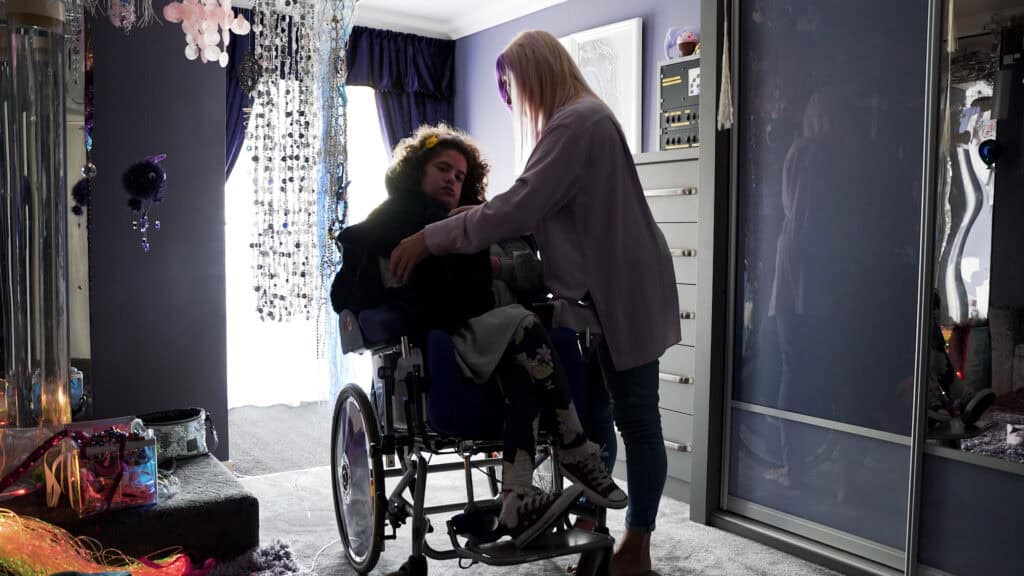 Yvette, a white woman with blonde and purple hair, putting a jacket on her daughter Rosey, a young woman with brown hair in a wheelchair.