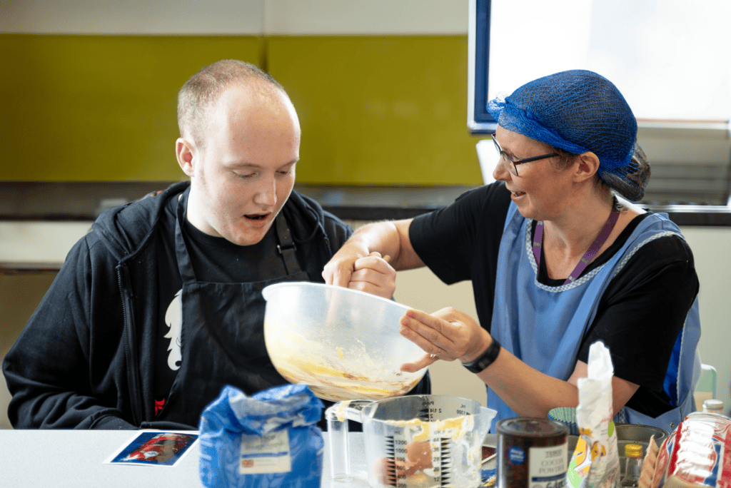 A young man sits next to his support worker, together they are mixing a bowl of cake batter.