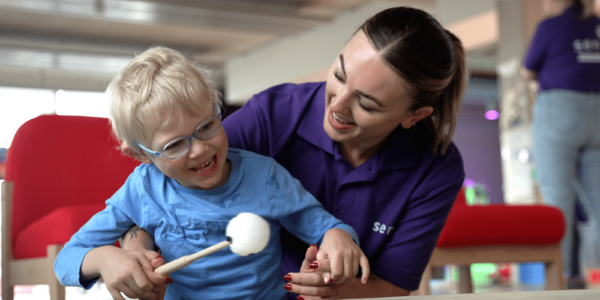 A young boy with blonde wavy hair and blue glasses grins as he holds a drum stick, about to bang a drum. A Sense support worker wearing a purple Sense polo shirt smiles at him and supports him to sit up.